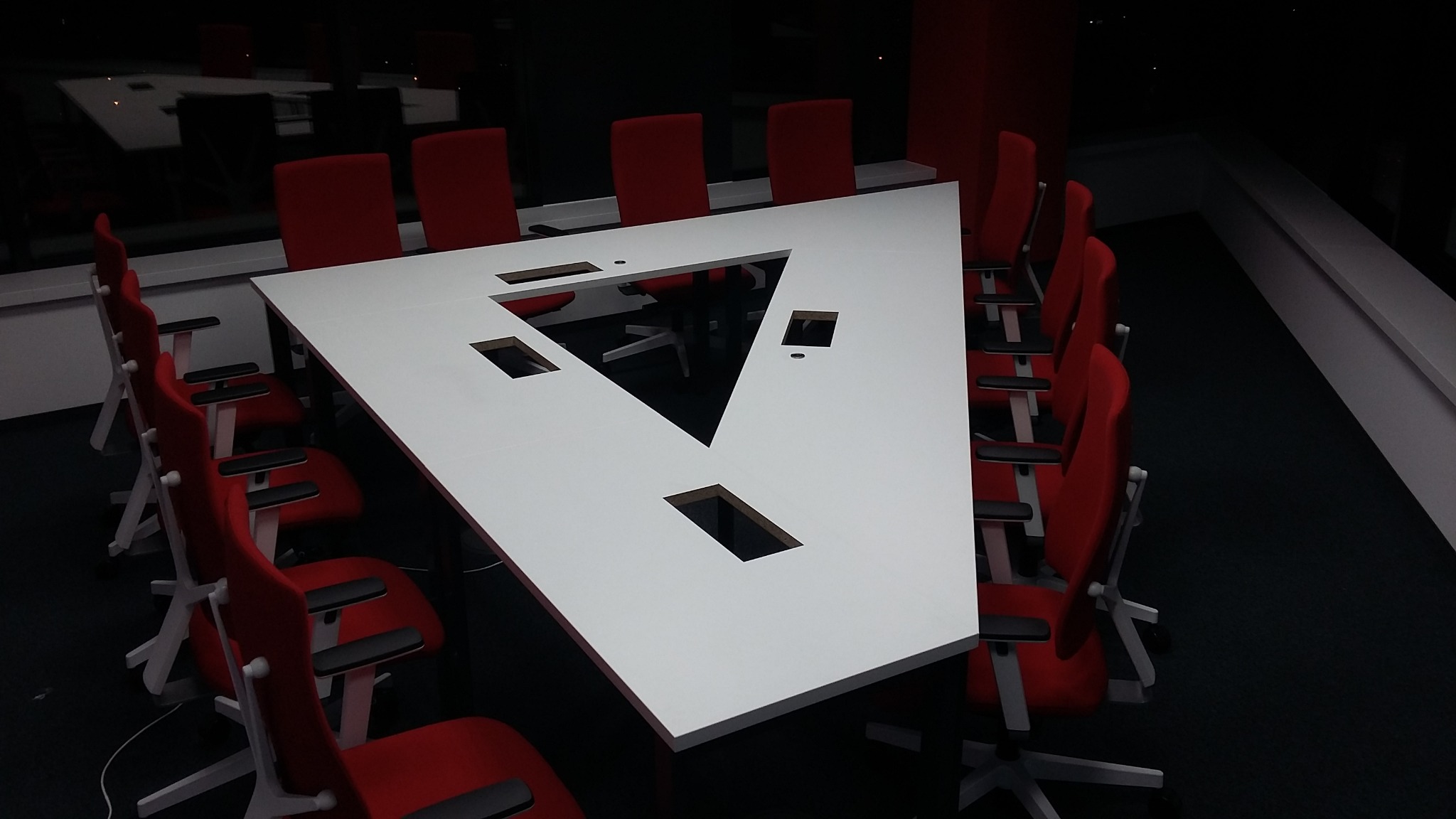 Nowoczesna sala konferencyjna z trójkątnym, białym stołem z otworami na kable i czerwonymi krzesłami na kółkach.