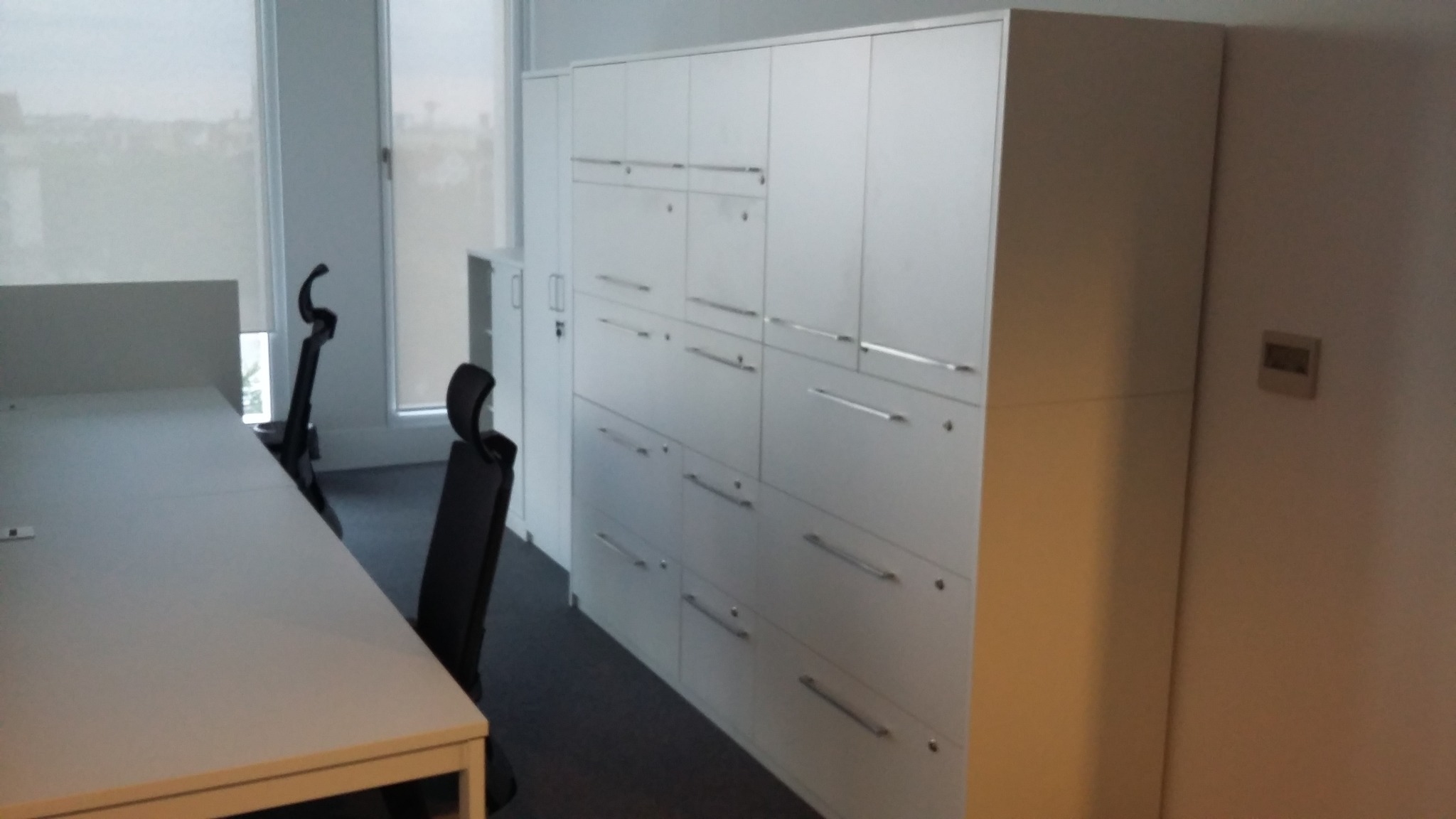 Białe szafy biurowe z szufladami i szafkami z metalowymi uchwytami, widoczne biurka i krzesła biurowe na tle okien z żaluzjami.