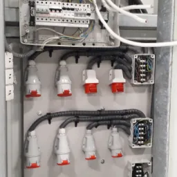 Rozdzielnia elektryczna z gniazdami siłowymi, widoczne przewody, zabezpieczenia i puszki przyłączeniowe.