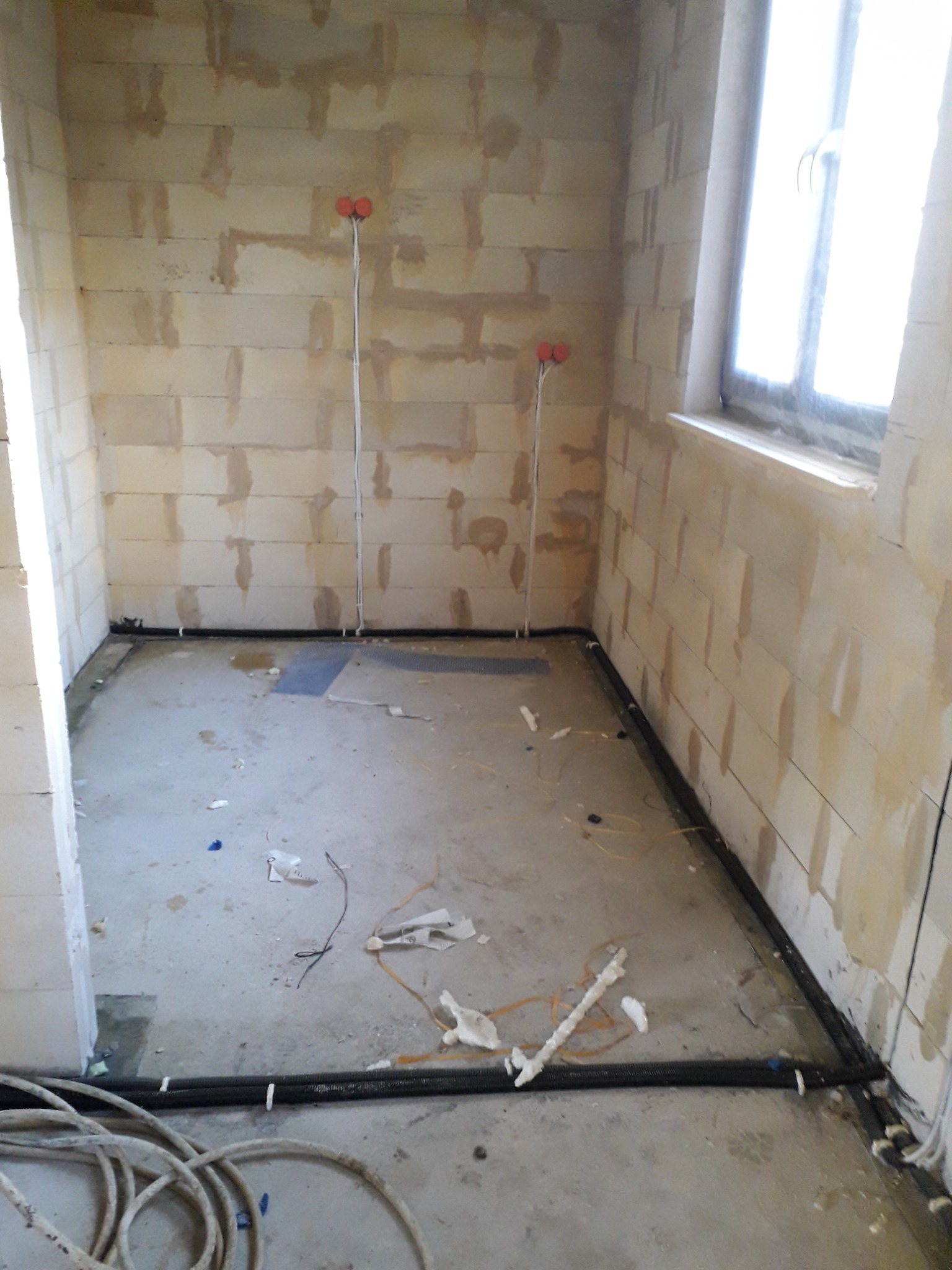 Surowy stan wnętrza w trakcie prac instalacyjnych: widoczne przewody elektryczne w peszlach ułożone wzdłuż ścian, puszki elektryczne na ścianach z betonu komórkowego, okno z jasnym światłem dziennym.