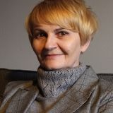 Portret kobiety z krótkimi blond włosami, ubranej w szary sweter i marynarkę w kratę, siedzącej na ciemnej sofie na tle szarej ściany.