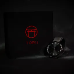 Zegarek Torii z czarną tarczą i bransoletą mesh, leżący obok czarnego pudełka z czerwonym logo firmy, sfotografowany w ciemnym otoczeniu.
