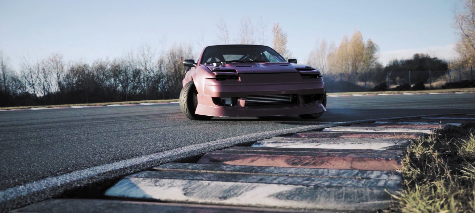 Przód bordowego samochodu driftowego na torze wyścigowym, widoczne detale karoserii i opony, tło stanowią drzewa i błękitne niebo.