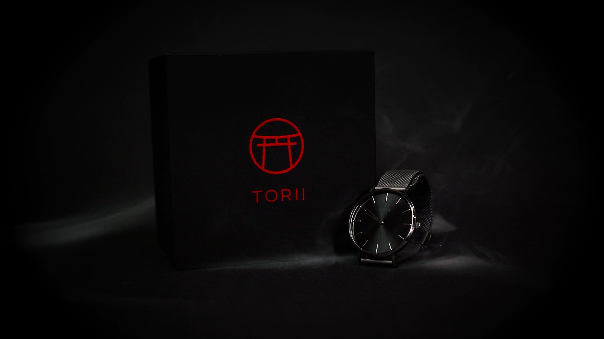 Zegarek Torii z czarną tarczą i bransoletą mesh, leżący obok czarnego pudełka z czerwonym logo firmy, sfotografowany w ciemnym otoczeniu.