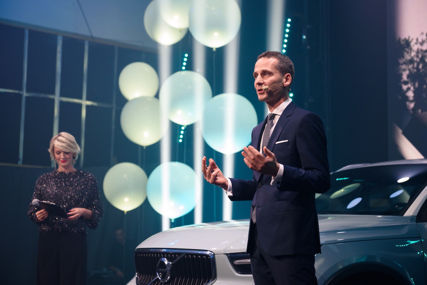 Prezentacja nowego Volvo: Mężczyzna w garniturze gestykuluje na scenie, obok stoi biały samochód, w tle balony i kobieta z mikrofonem.