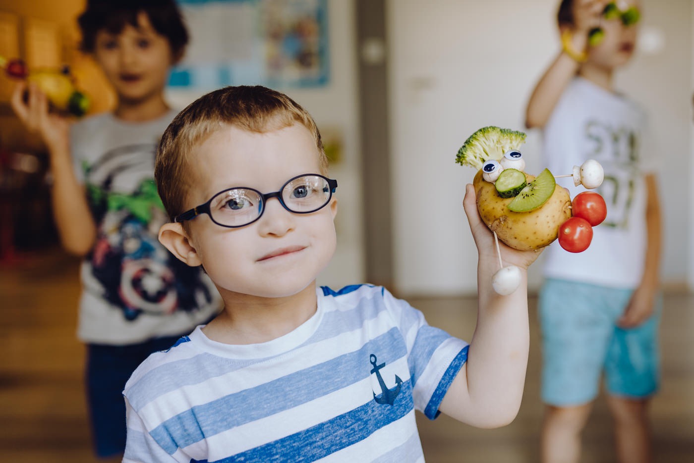 Chłopiec w okularach trzyma ziemniaczaną figurkę z brokułami, kiwi, pomidorami i pieczarkami, wykonaną na warsztatach kulinarnych dla dzieci.