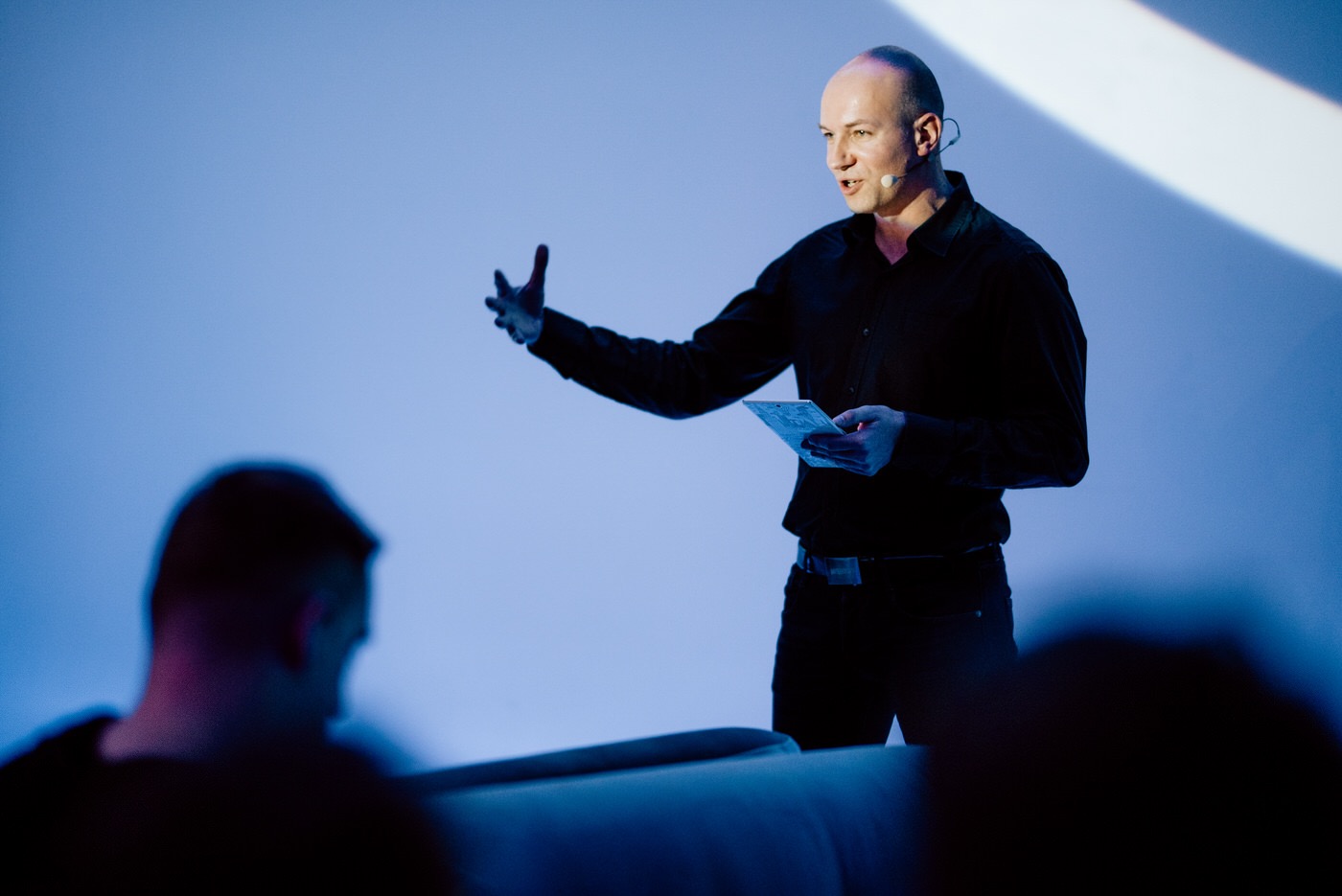 Mężczyzna w czarnej koszuli i spodniach, z mikrofonem nagłownym, gestykuluje podczas prezentacji na tle niebieskiego ekranu, trzymając w dłoniach tablet.