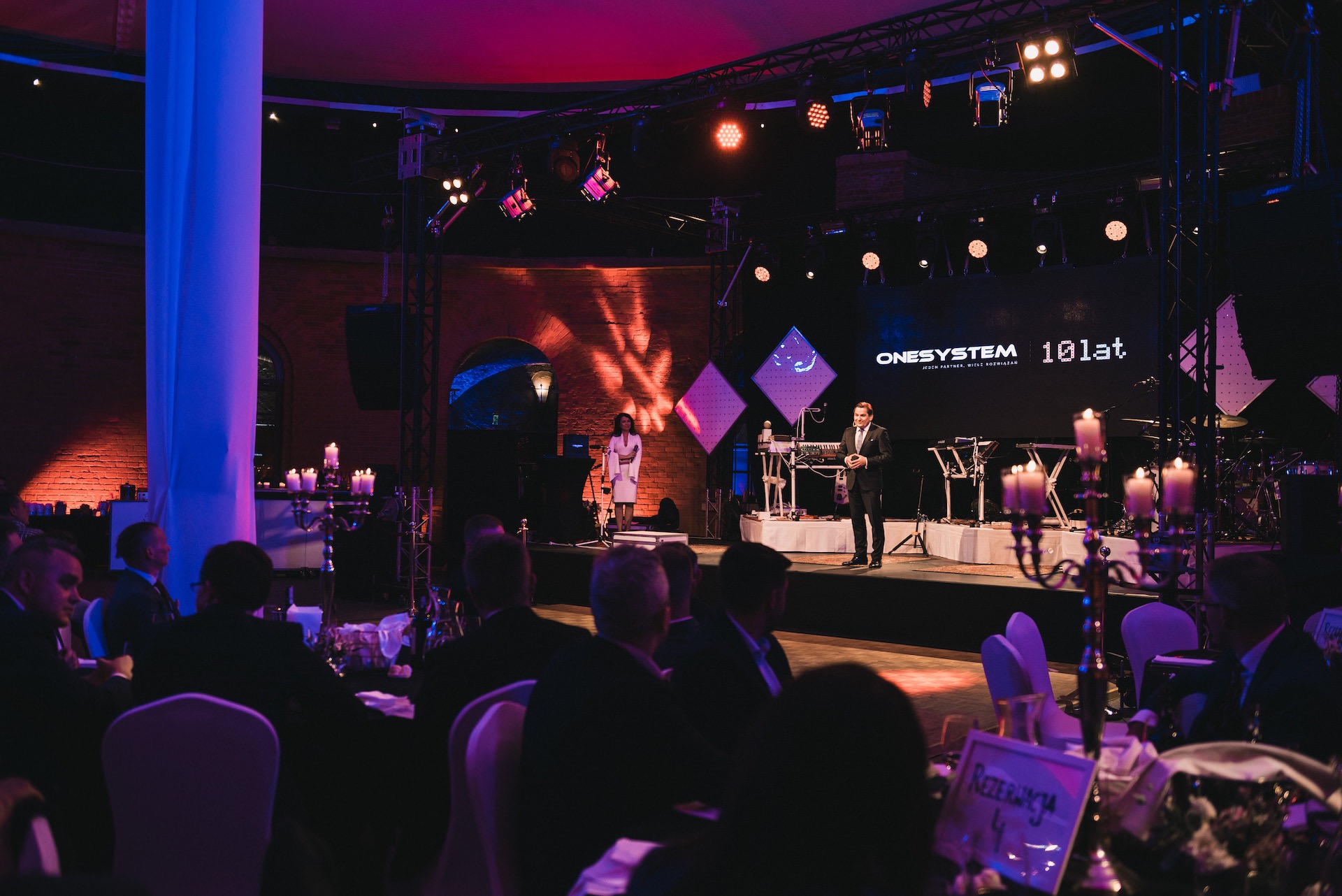 Uroczysta gala: scena z oświetleniem, występujący konferansjer w garniturze, w tle wokalistka i ekran z logo firmy, na pierwszym planie stoliki z gośćmi i świeczniki.