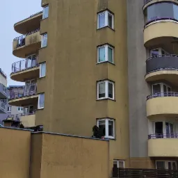 Wielopiętrowy budynek z balkonami i elewacją w odcieniach beżu i szarości, z widocznymi śladami zabrudzeń na ścianach, zieleń i śmieci na trawniku.