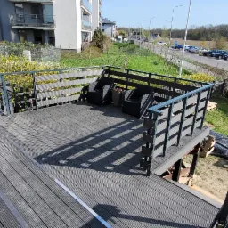 Nowoczesny taras z kompozytowych desek w odcieniach szarości, z metalową balustradą, częściowo zadaszony, z widocznymi pracami wykończeniowymi i elementami wyposażenia.