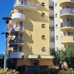 Żółto-biały budynek mieszkalny z zaokrąglonymi balkonami i okrągłymi oknami, widziany z poziomu ulicy w słoneczny dzień.