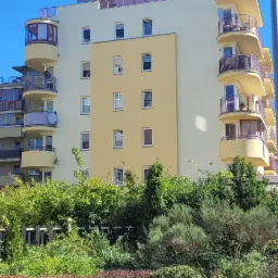 Wielopiętrowy budynek mieszkalny z zaokrąglonymi balkonami i elewacją w odcieniach żółci, częściowo zasłonięty zielenią.