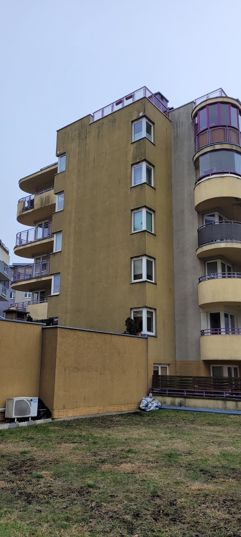 Wielopiętrowy budynek z balkonami i elewacją w odcieniach beżu i szarości, z widocznymi śladami zabrudzeń na ścianach, zieleń i śmieci na trawniku.