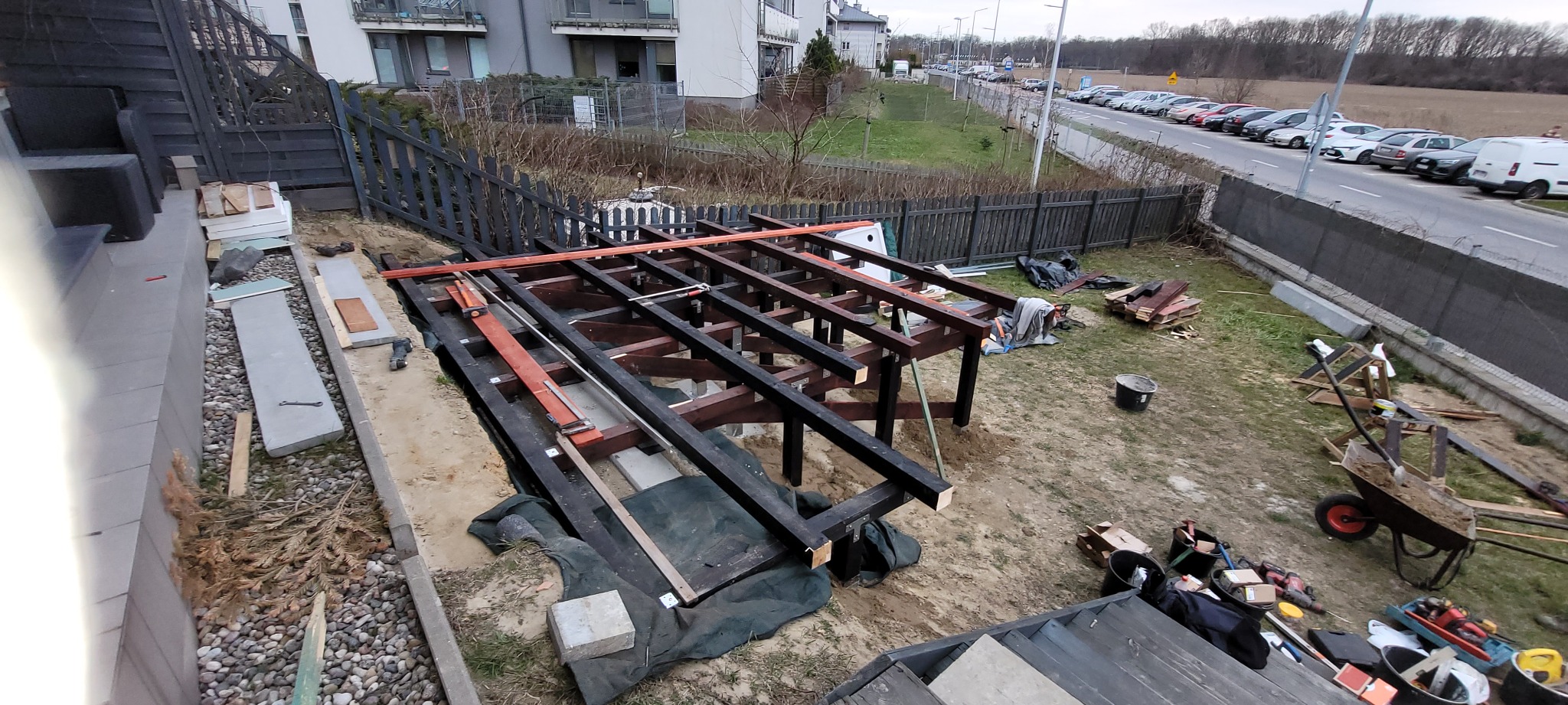 Konstrukcja nośna tarasu z brązowych belek drewnianych, przygotowana do montażu deski kompozytowej, widoczna z góry na tle trawnika i zabudowy wielorodzinnej.