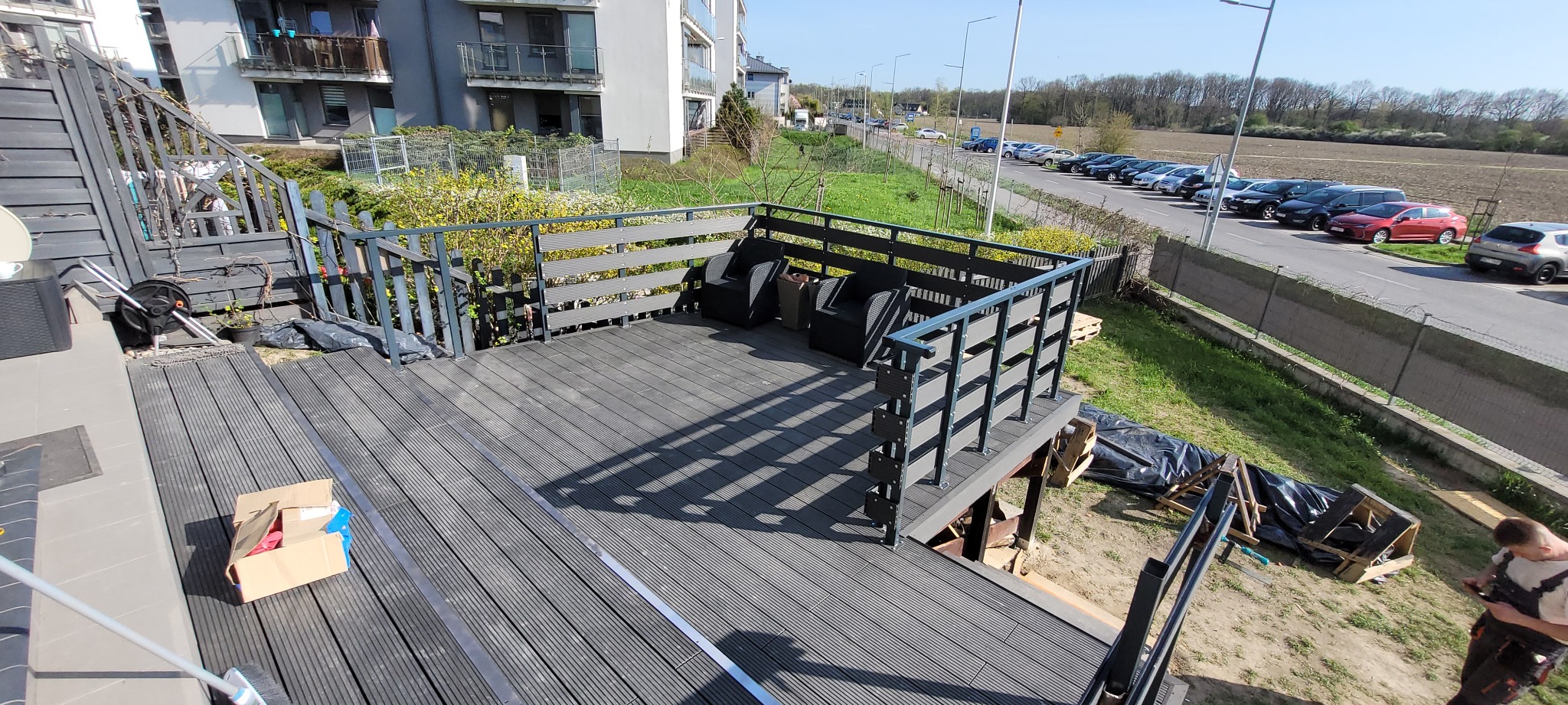 Nowoczesny taras z kompozytowych desek w odcieniach szarości, z metalową balustradą, częściowo zadaszony, z widocznymi pracami wykończeniowymi i elementami wyposażenia.