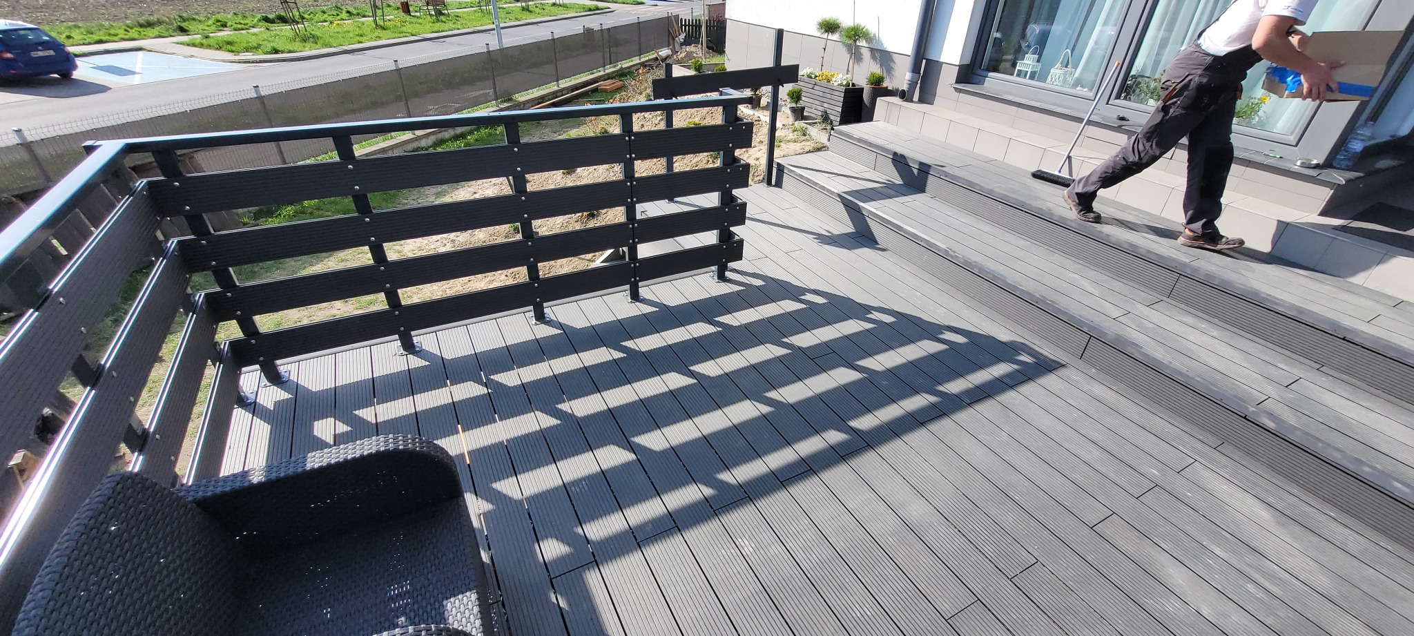 Szara, kompozytowa deska tarasowa z czarną, poziomą balustradą i fragmentem fotela z technorattanu, widok z góry na fragment domu z oknem i osobą w kombinezonie niosącą karton.