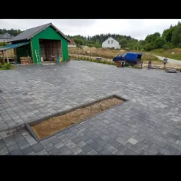 Szeroki, szary podjazd z kostki brukowej w Gdańsku, z wydzielonym miejscem na rośliny wypełnionym piaskiem, w tle zielony garaż i dom w budowie.