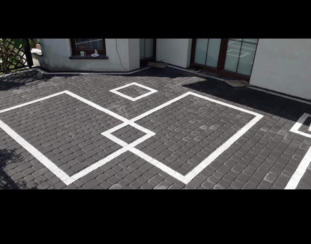 Szara kostka brukowa ułożona w geometryczny wzór z białymi kwadratami przed budynkiem z oknami.