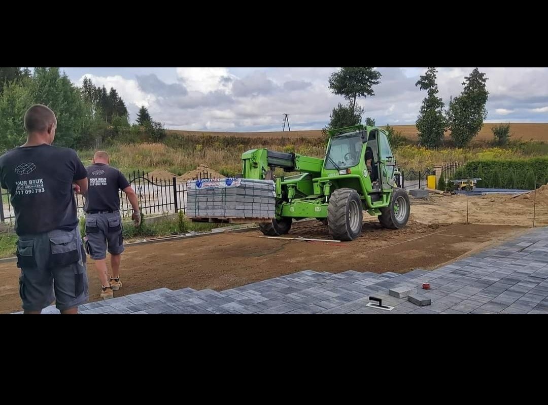 Zielona ładowarka teleskopowa transportuje paletę z kostką brukową na placu budowy, gdzie dwóch pracowników w koszulkach z logo firmy nadzoruje prace, a w tle widoczne jest ogrodzenie i krajobraz...