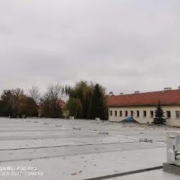 Widok z góry na płaski dach pokryty panelami, z zamontowanymi jednostkami zewnętrznymi klimatyzacji, z budynkiem w tle i jesiennymi drzewami, pochmurne niebo.