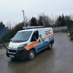 Biały samochód dostawczy z grafiką i danymi kontaktowymi firmy oferującej serwis klimatyzacji i AGD, zaparkowany na mokrym podjeździe z kostki brukowej, drabina na dachu.