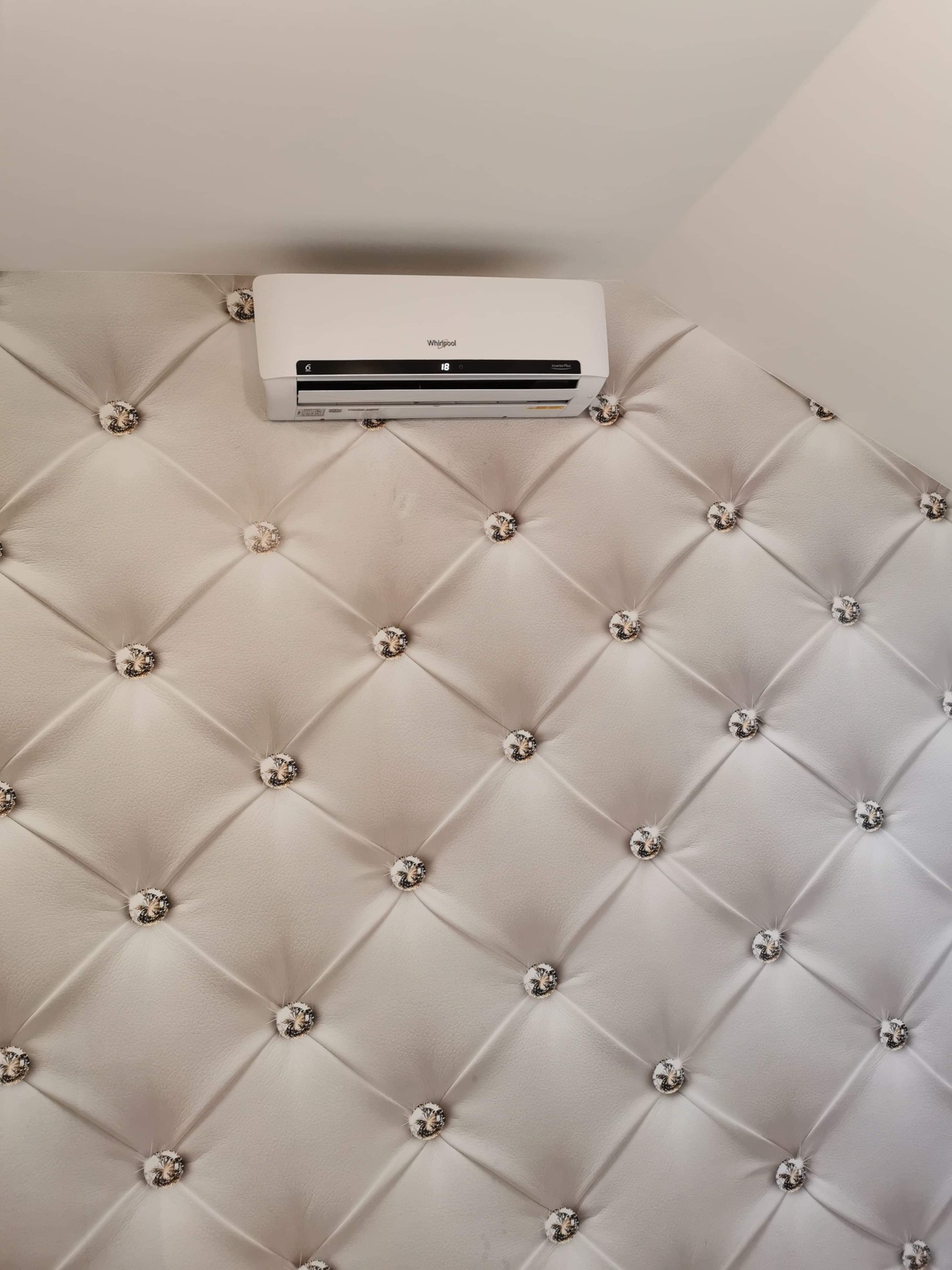 Biały klimatyzator ścienny marki Whirlpool zamontowany na ozdobnej, pikowanej ścianie z dekoracyjnymi kryształkami.
