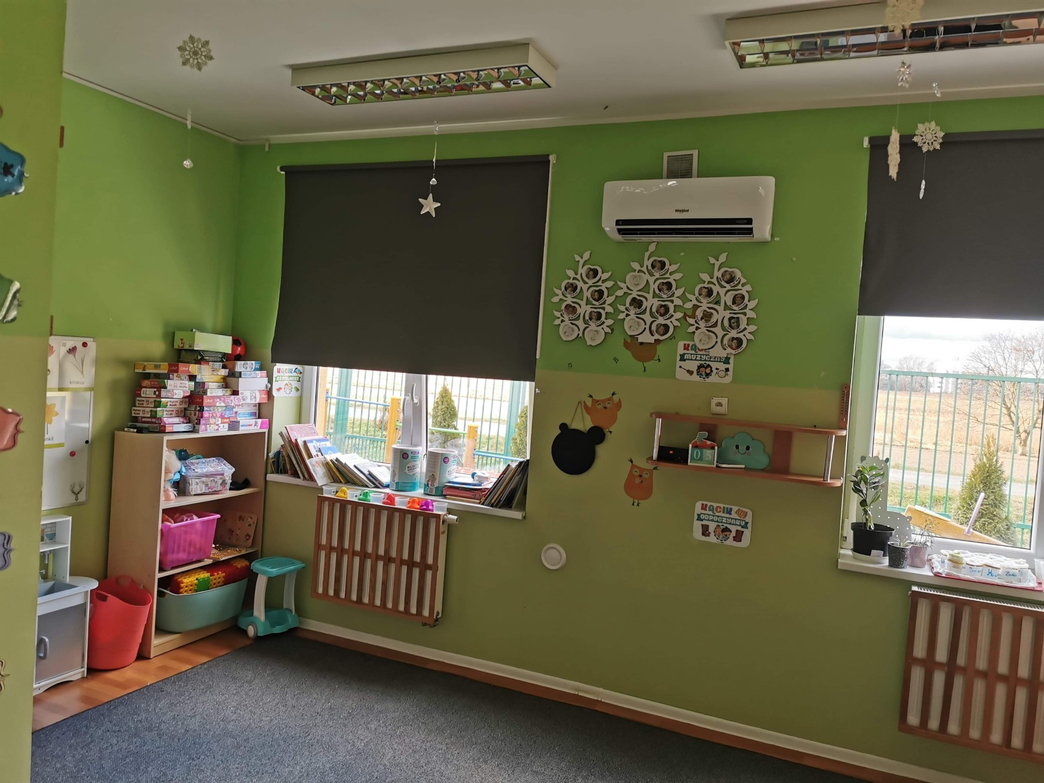 Wnętrze przedszkolnej sali z zielonymi ścianami, grzejnikami pod oknami, regałem z zabawkami i klimatyzatorem Whirlpool na ścianie.