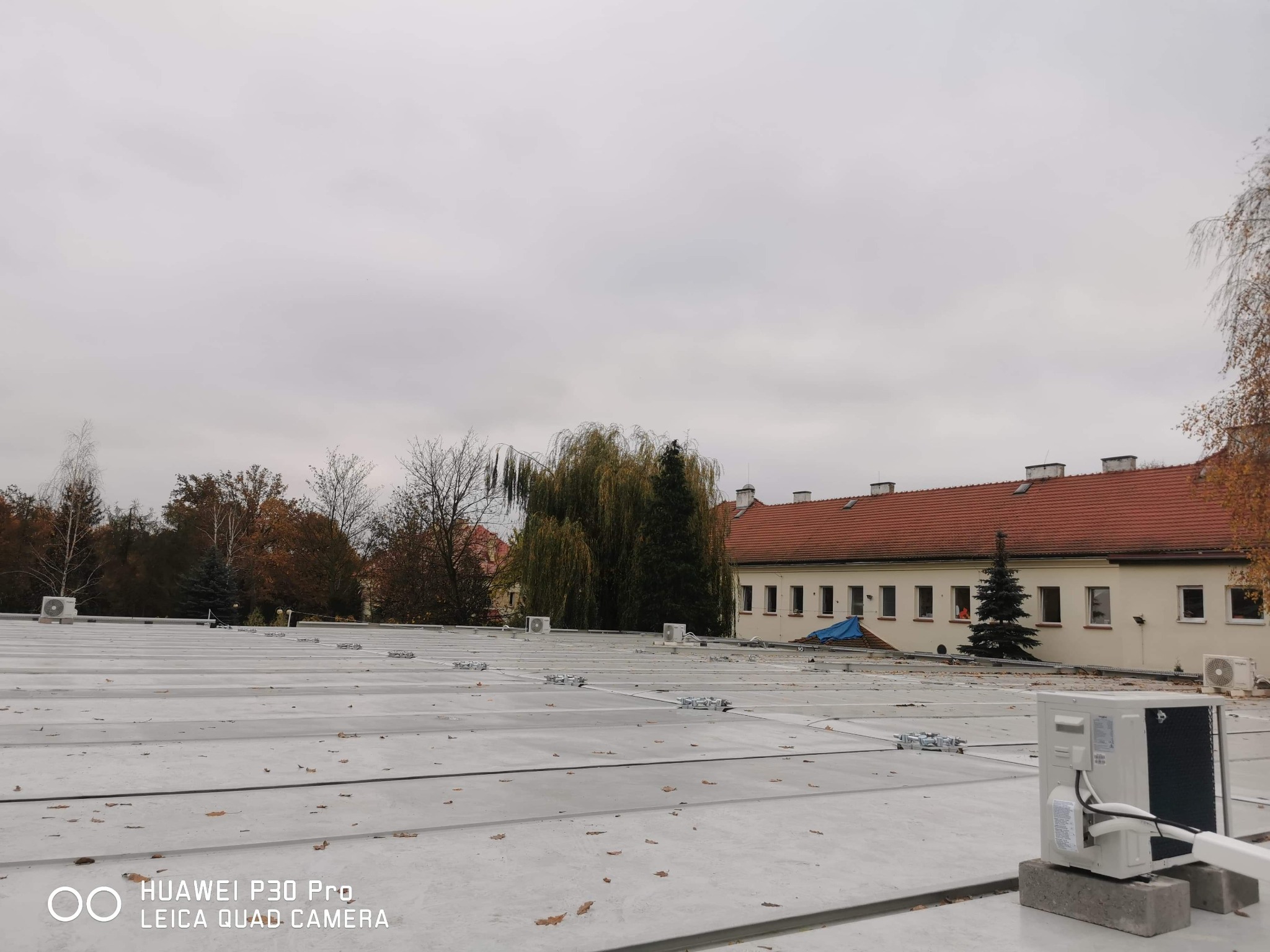 Widok z góry na płaski dach pokryty panelami, z zamontowanymi jednostkami zewnętrznymi klimatyzacji, z budynkiem w tle i jesiennymi drzewami, pochmurne niebo.
