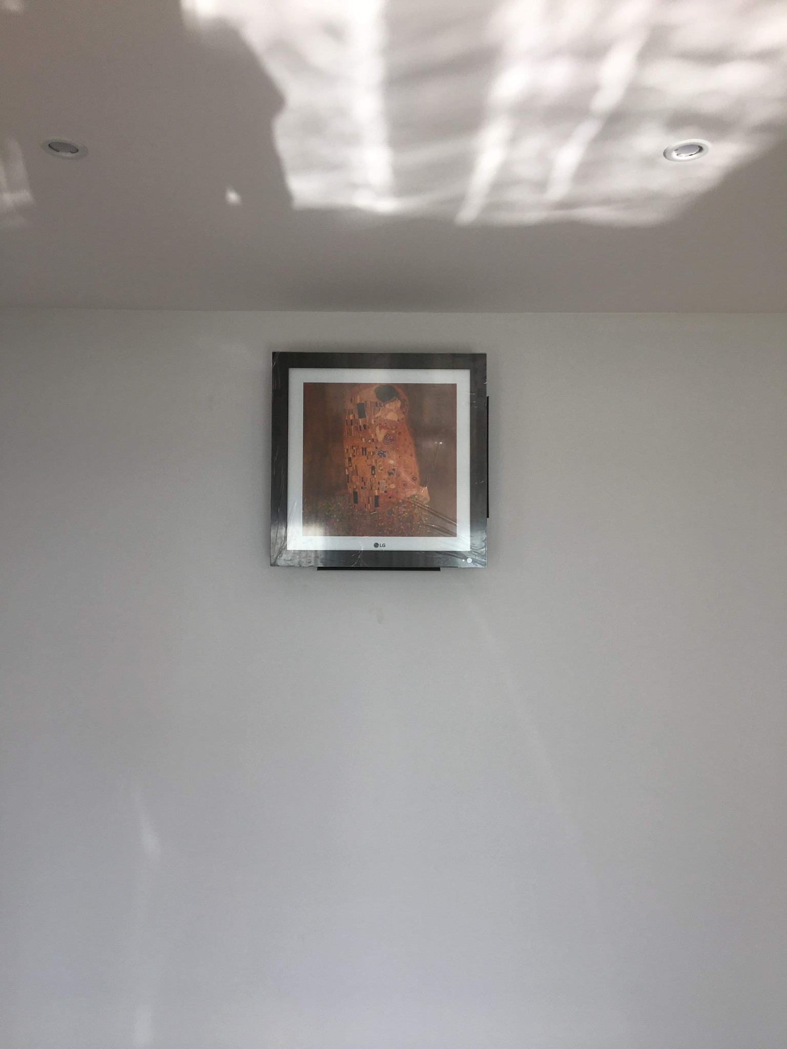 Obraz Gustava Klimta 'Pocałunek' w ramie, zawieszony na szarej ścianie pod sufitem z wbudowanymi lampami, widoczne odbicie światła na suficie.