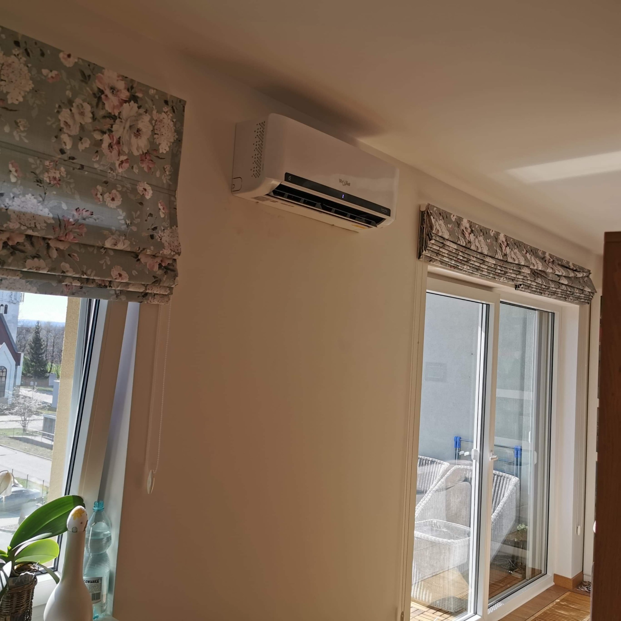 Biała jednostka klimatyzacji zamontowana na ścianie w jasnym pokoju z oknami zasłoniętymi roletami rzymskimi w kwiatowy wzór. Widok na balkon z białymi meblami.
