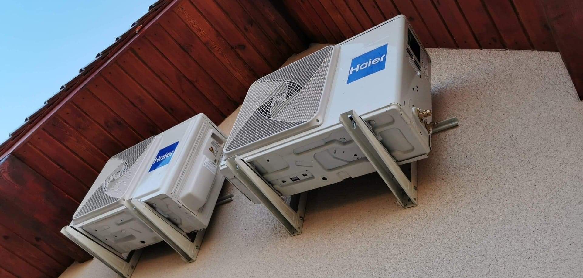 Dwa białe klimatyzatory Haier zamontowane na zewnątrz budynku z beżową elewacją i drewnianym okapem.
