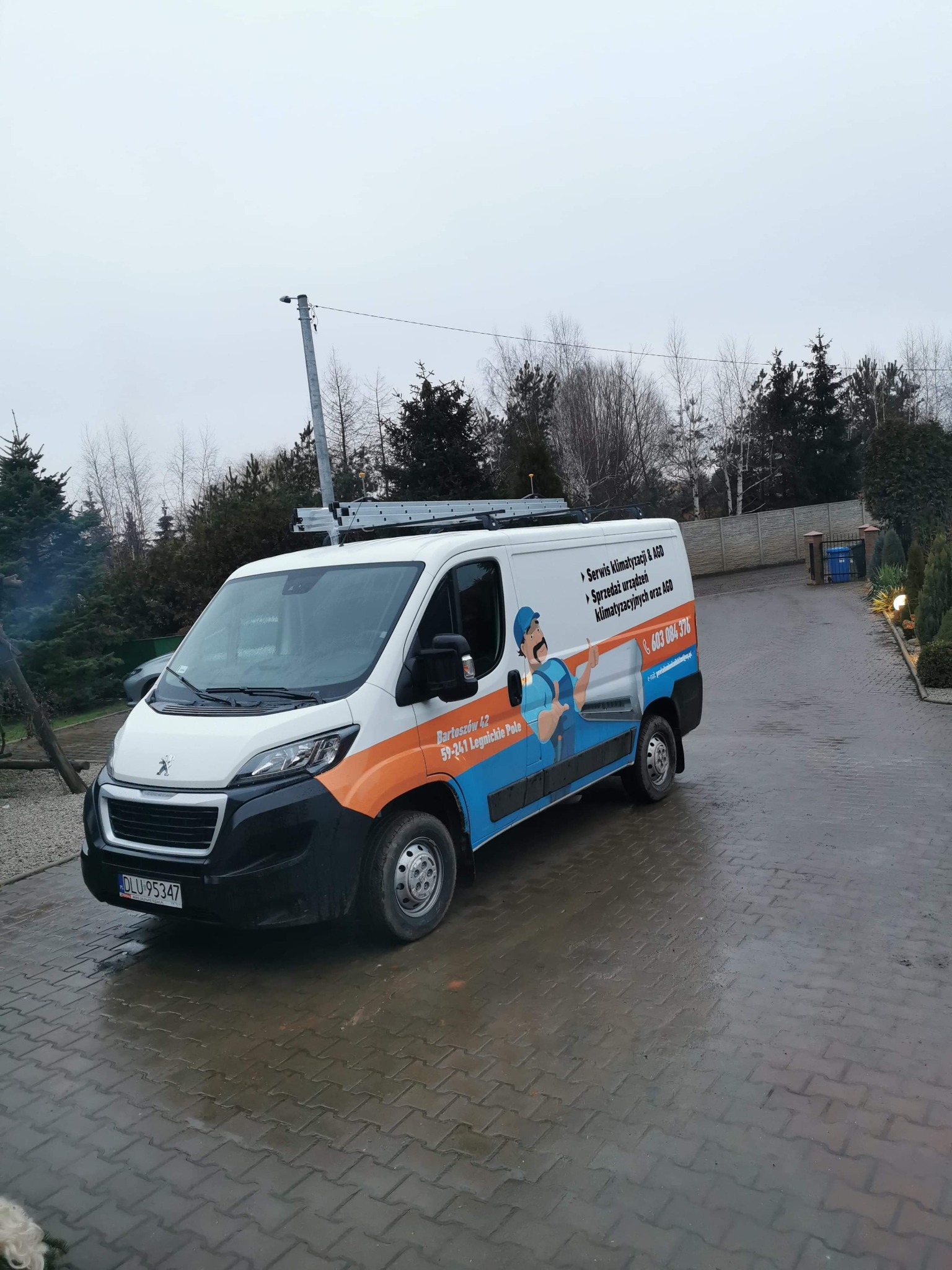 Biały samochód dostawczy z grafiką i danymi kontaktowymi firmy oferującej serwis klimatyzacji i AGD, zaparkowany na mokrym podjeździe z kostki brukowej, drabina na dachu.