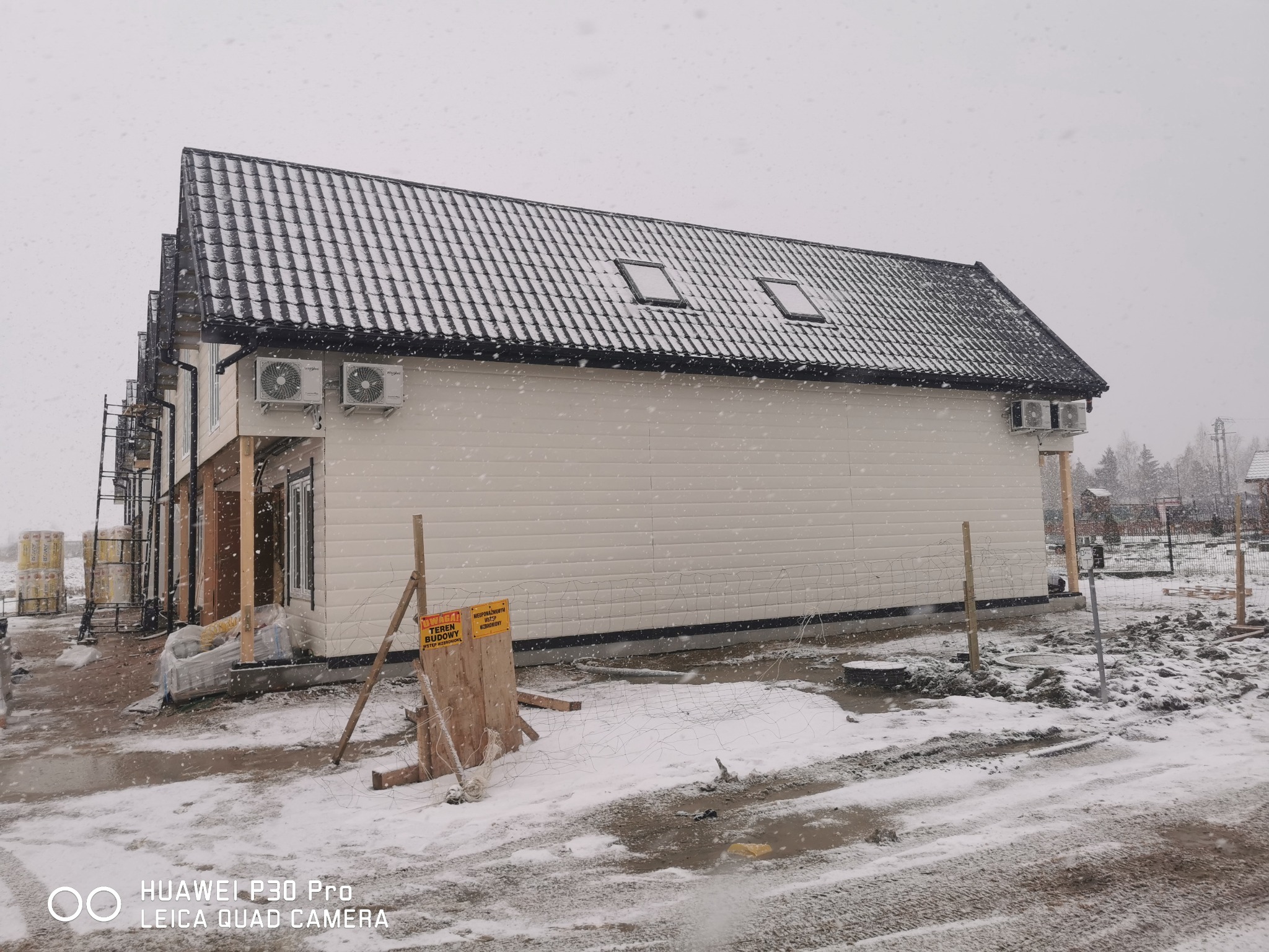 Budynek w trakcie budowy z zamontowanymi klimatyzatorami zewnętrznymi na ścianie, pokryty śniegiem, widoczne oznakowanie terenu budowy.