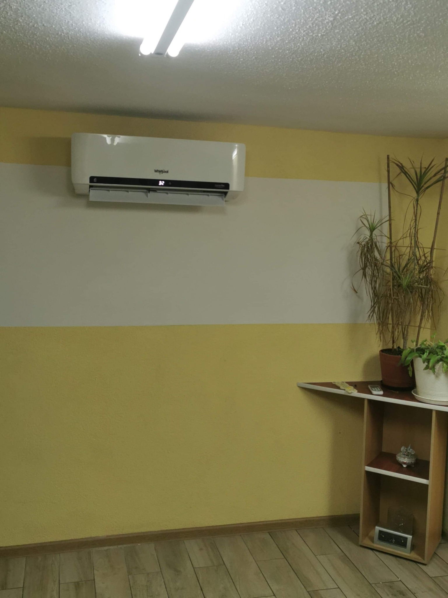 Biały klimatyzator marki Whirlpool zamontowany na ścianie pomalowanej na żółto i biało, w pomieszczeniu z drewnopodobną podłogą i półką z roślinami.