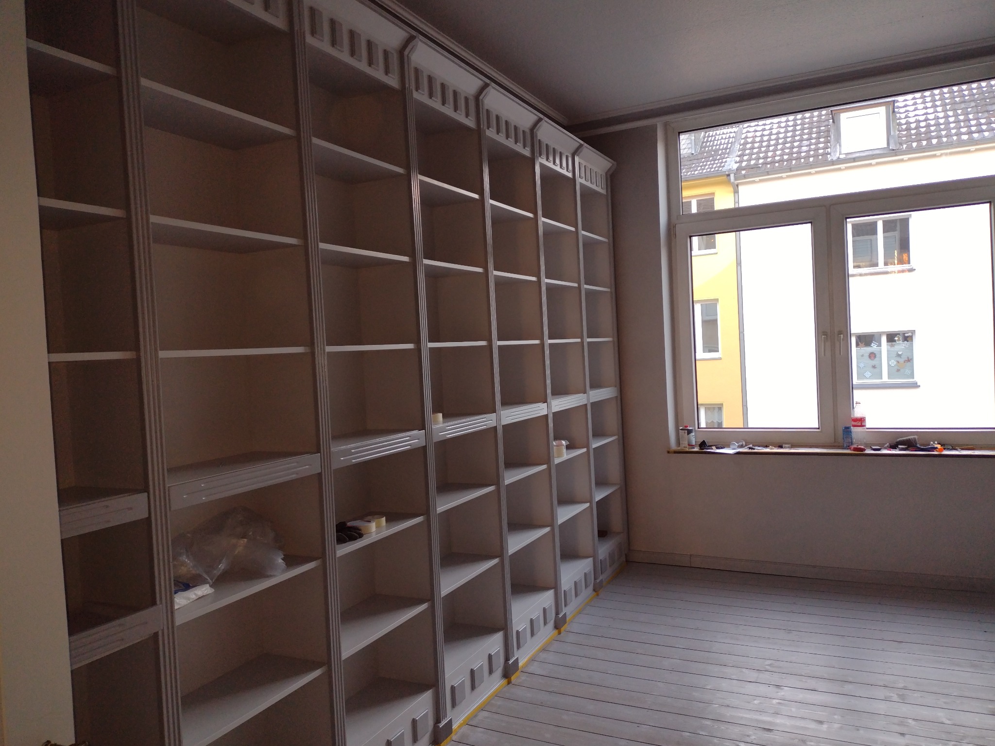 Białe, klasyczne regały w trakcie montażu, zabezpieczone taśmą malarską, widok na szare, pomalowane podłogi i okno z widokiem na budynki