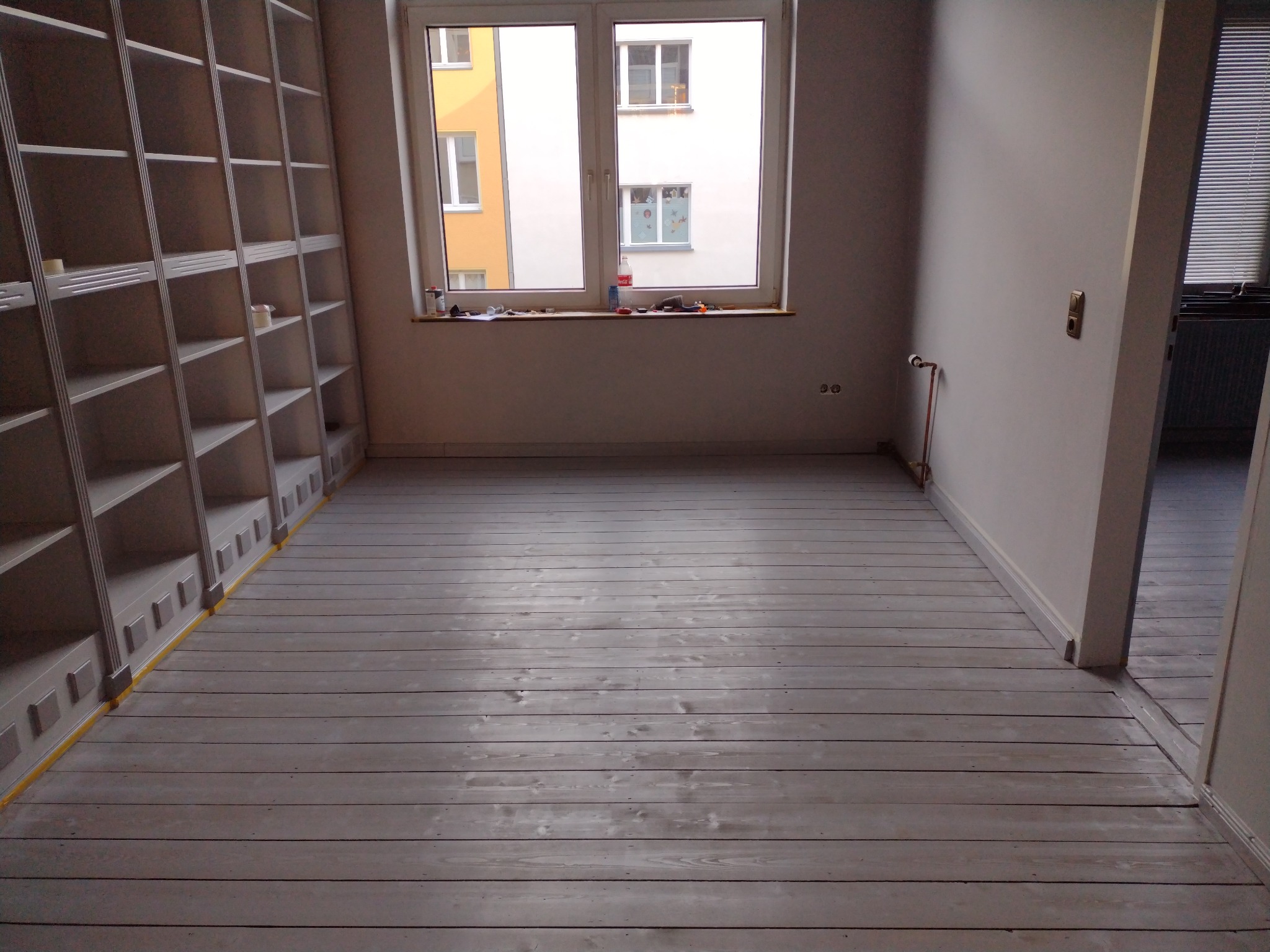 Pokój po malowaniu, z pomalowaną na biało drewnianą podłogą, wbudowanym regałem na ścianie i oknem z widokiem na budynki