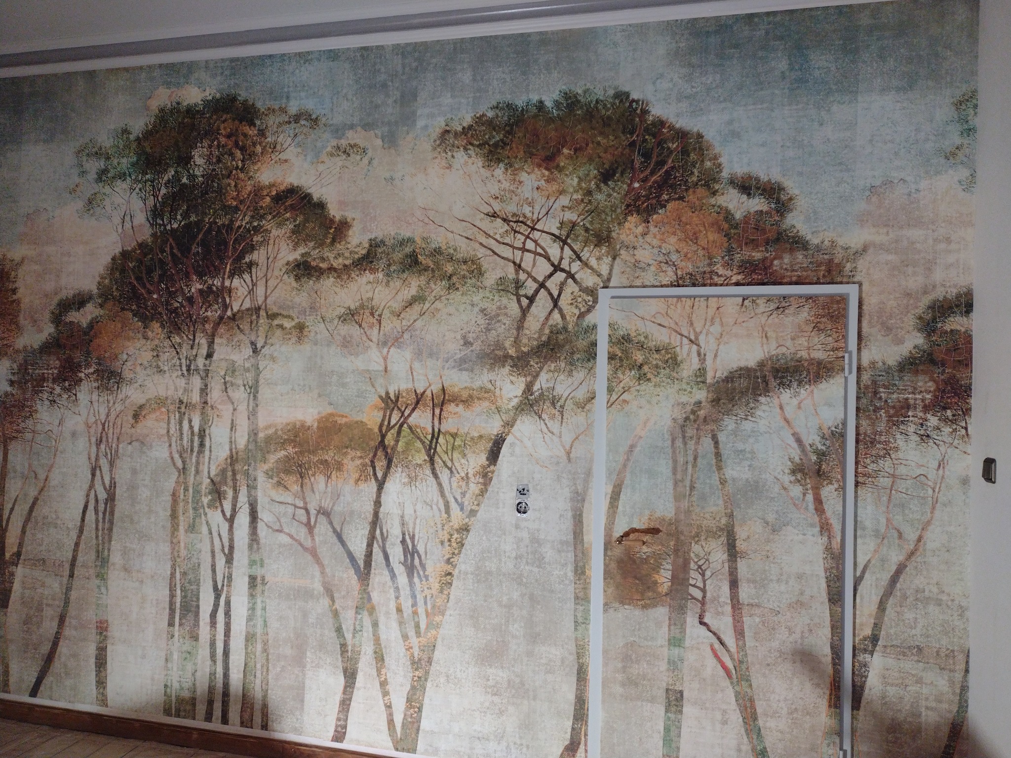 Fototapeta z motywem drzew w stylu malarskim zdobiącą ścianę w pokoju, widoczna rama drzwi i gniazdka elektryczne.