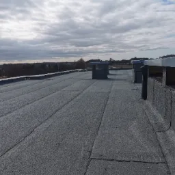 Płaski dach pokryty papą termozgrzewalną z widocznymi kominkami wentylacyjnymi i obróbkami blacharskimi na tle pochmurnego nieba i zabudowań.