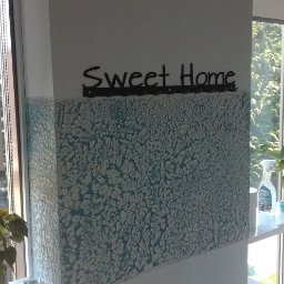 Usługi remontowe Warszawa i okolice - Fragment ściany z dekoracyjną, błękitno-białą fakturą, nad którą umieszczono napis 'Sweet Home' na czarnym wieszaku, widoczne okno z roślinami w tle.