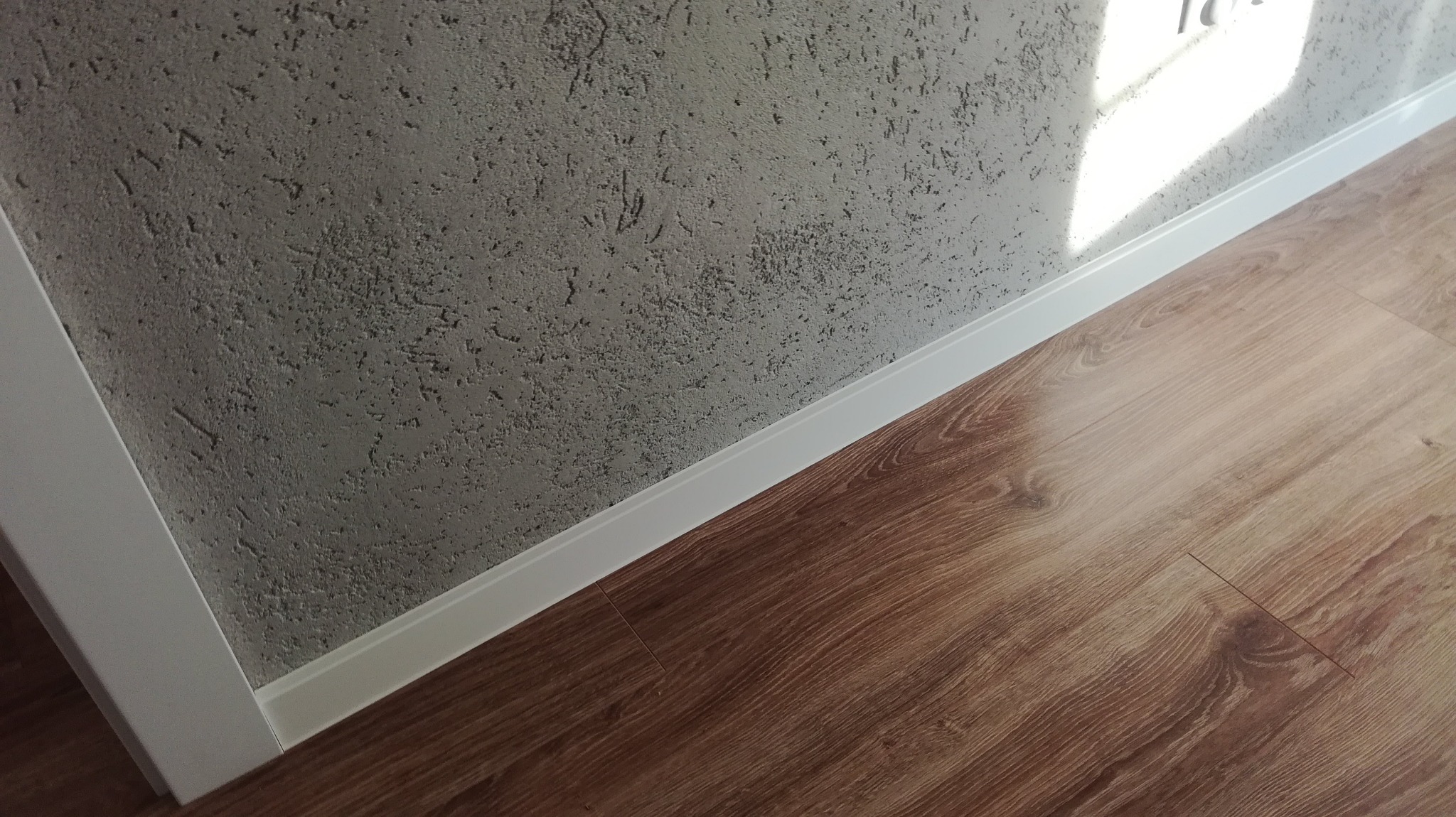 Szara ściana z efektem dekoracyjnym, biała listwa przypodłogowa i panele podłogowe imitujące drewno, oświetlone padającym światłem słonecznym.