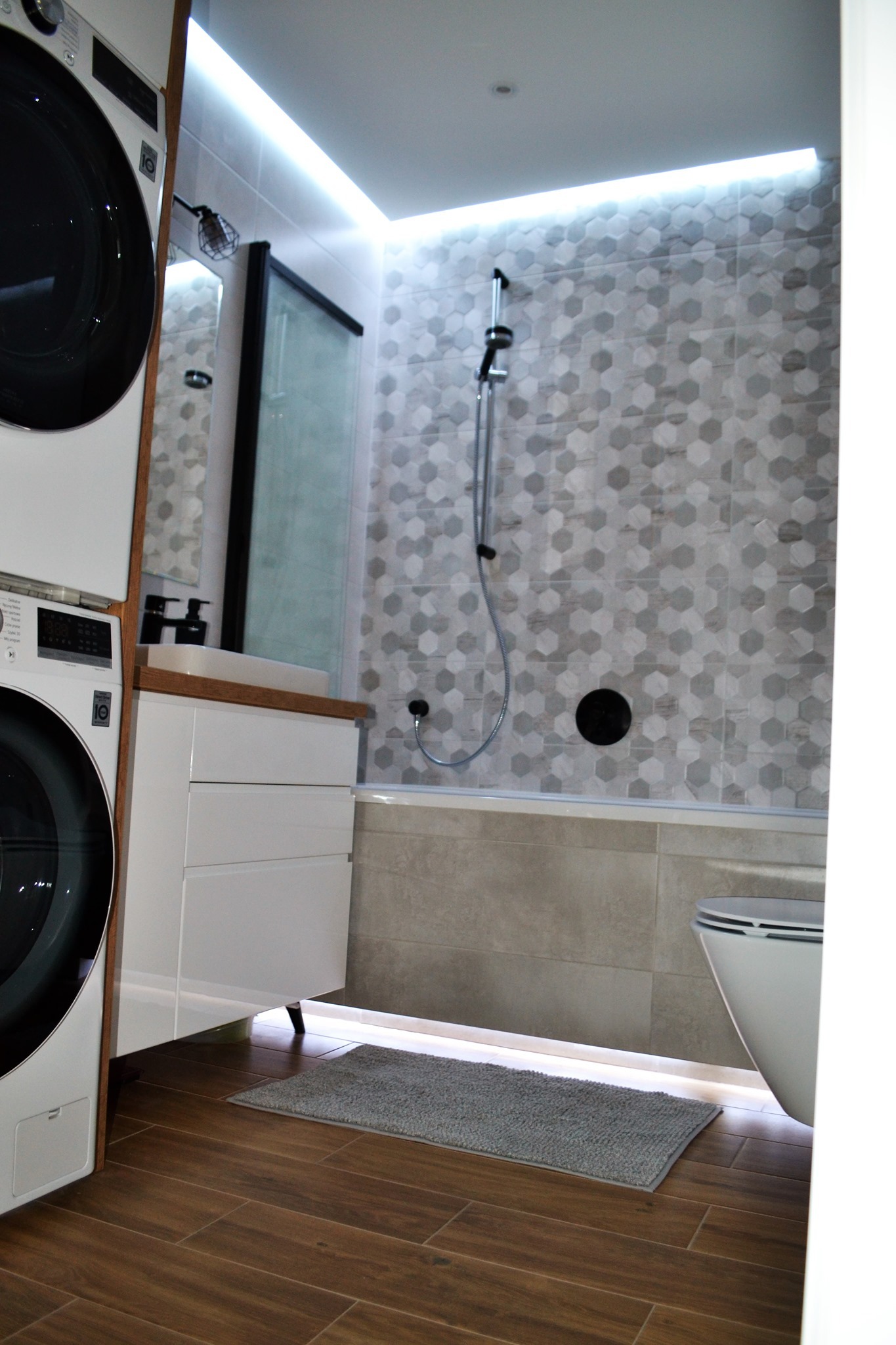 Nowoczesna łazienka z pralką i suszarką w zabudowie, heksagonalne płytki na ścianie prysznica, oświetlenie LED podsufitowe i pod wanną, drewnopodobna podłoga.