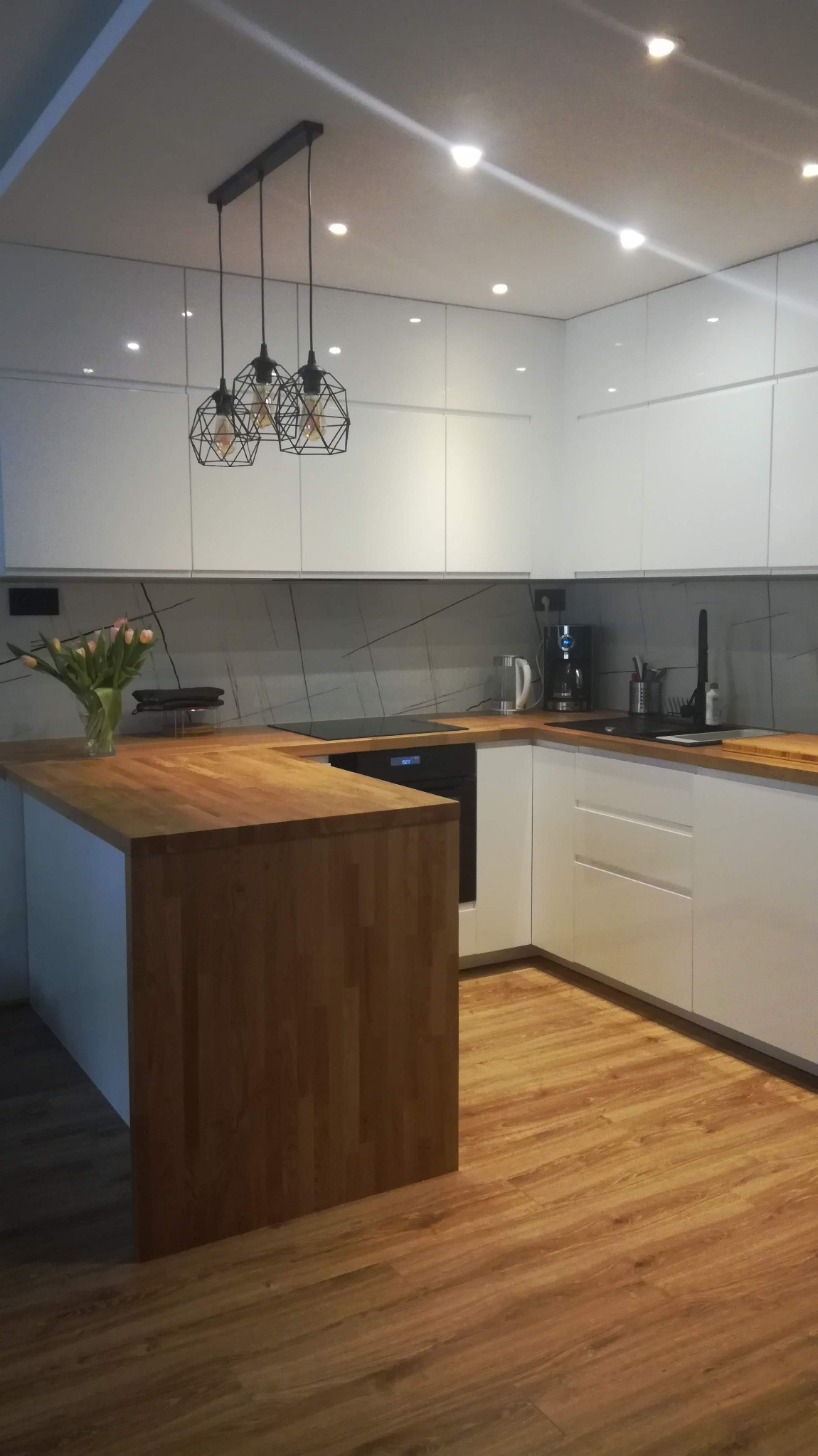 Nowoczesna kuchnia z białymi, błyszczącymi szafkami, drewnianym blatem i wyspą, oświetlona trzema geometrycznymi lampami wiszącymi i punktowymi światłami w suficie podwieszanym. Na blacie stoi...