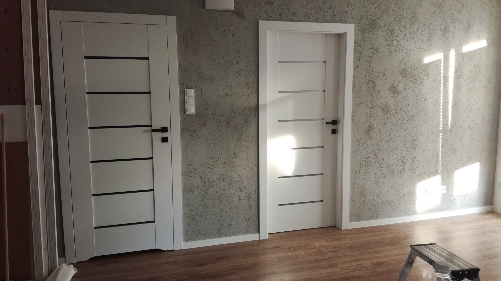 Dwa białe, nowoczesne drzwi wewnętrzne z czarnymi poziomymi wstawkami w nowym mieszkaniu. Na ścianie widoczna faktura tynku dekoracyjnego w odcieniach szarości. Na podłodze panele drewnopodobne.