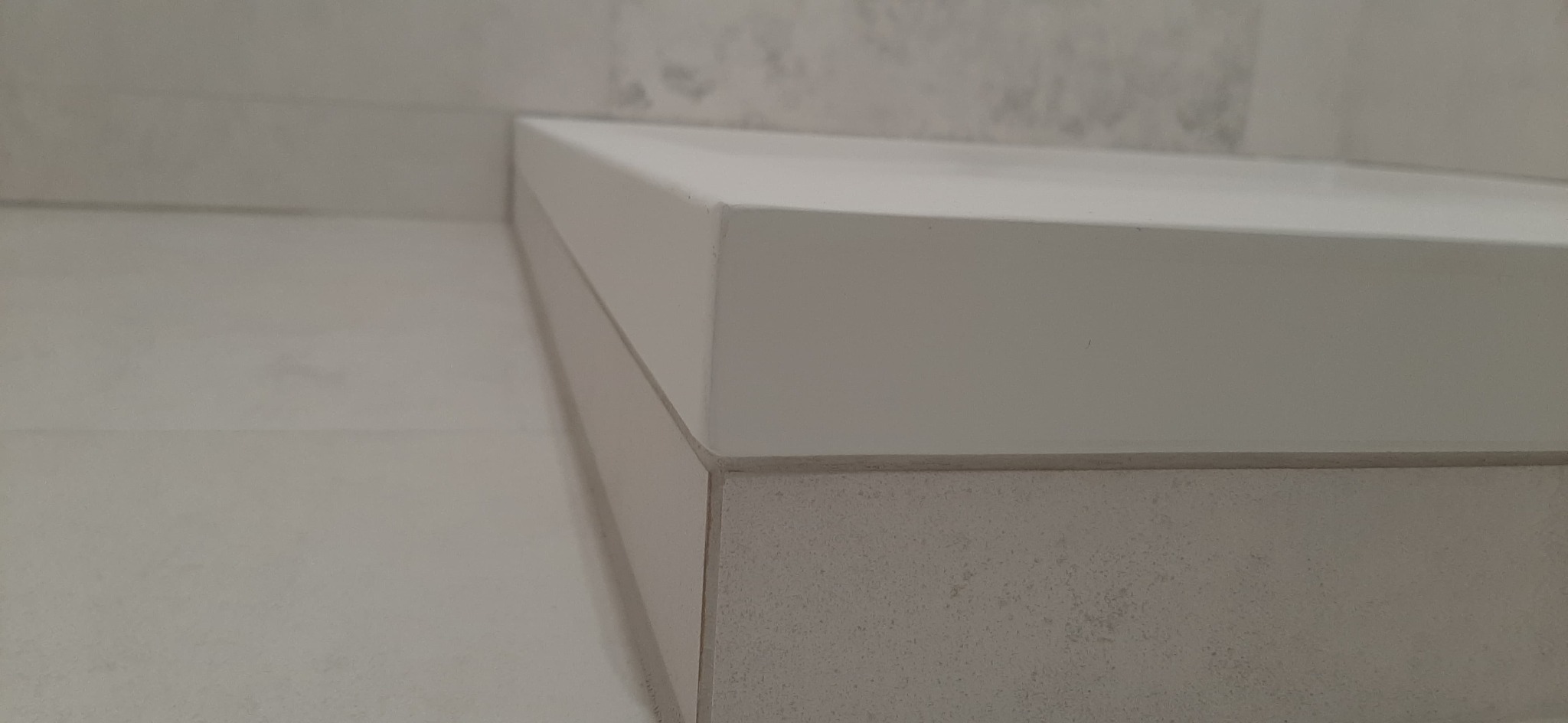 Biały blat umywalki z zaokrąglonymi krawędziami, dopasowany kolorystycznie do płytek ściennych i podłogowych w łazience, z widocznymi detalami łączenia materiałów.