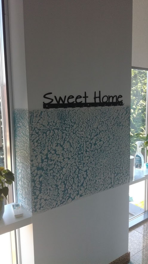 Fragment ściany z dekoracyjną, błękitno-białą fakturą, nad którą umieszczono napis 'Sweet Home' na czarnym wieszaku, widoczne okno z roślinami w tle.