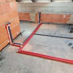 Wnętrze w trakcie budowy z widocznymi, częściowo zamontowanymi rurami instalacji hydraulicznej i grzewczej w kolorach czerwonym, białym i niebieskim, wystającymi ze ściany z cegły dziurawki oraz...