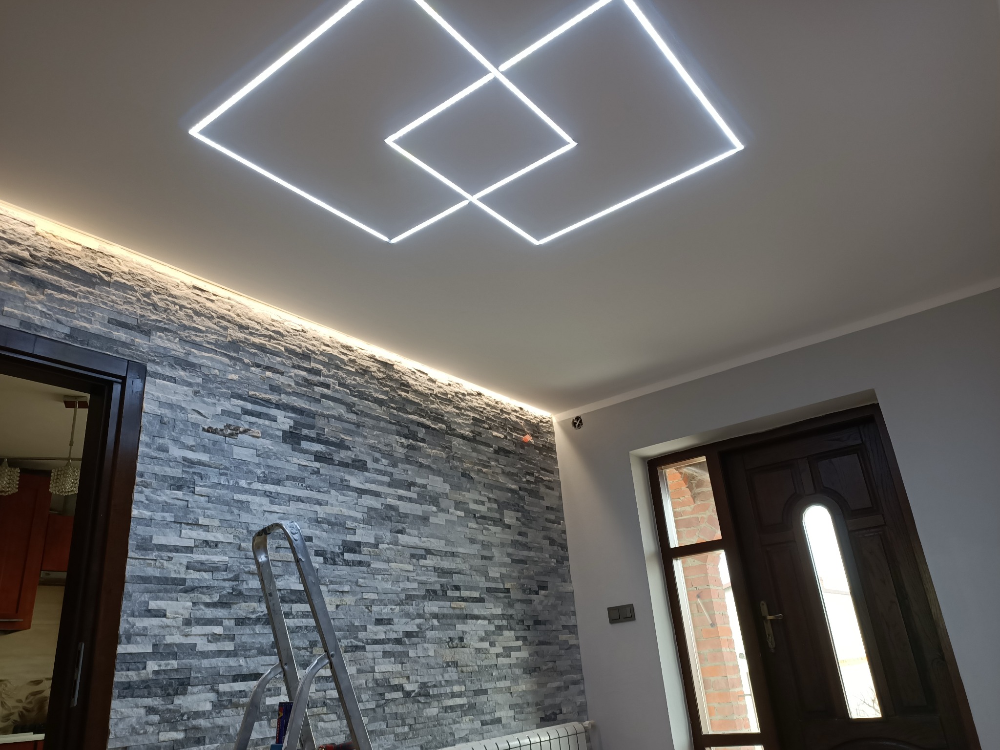 Nowoczesne oświetlenie sufitowe LED w kształcie dwóch kwadratów, ściana wykończona szarą okładziną imitującą kamień, drabina oparta o ścianę, widoczna część grzejnika i drzwi w tle.
