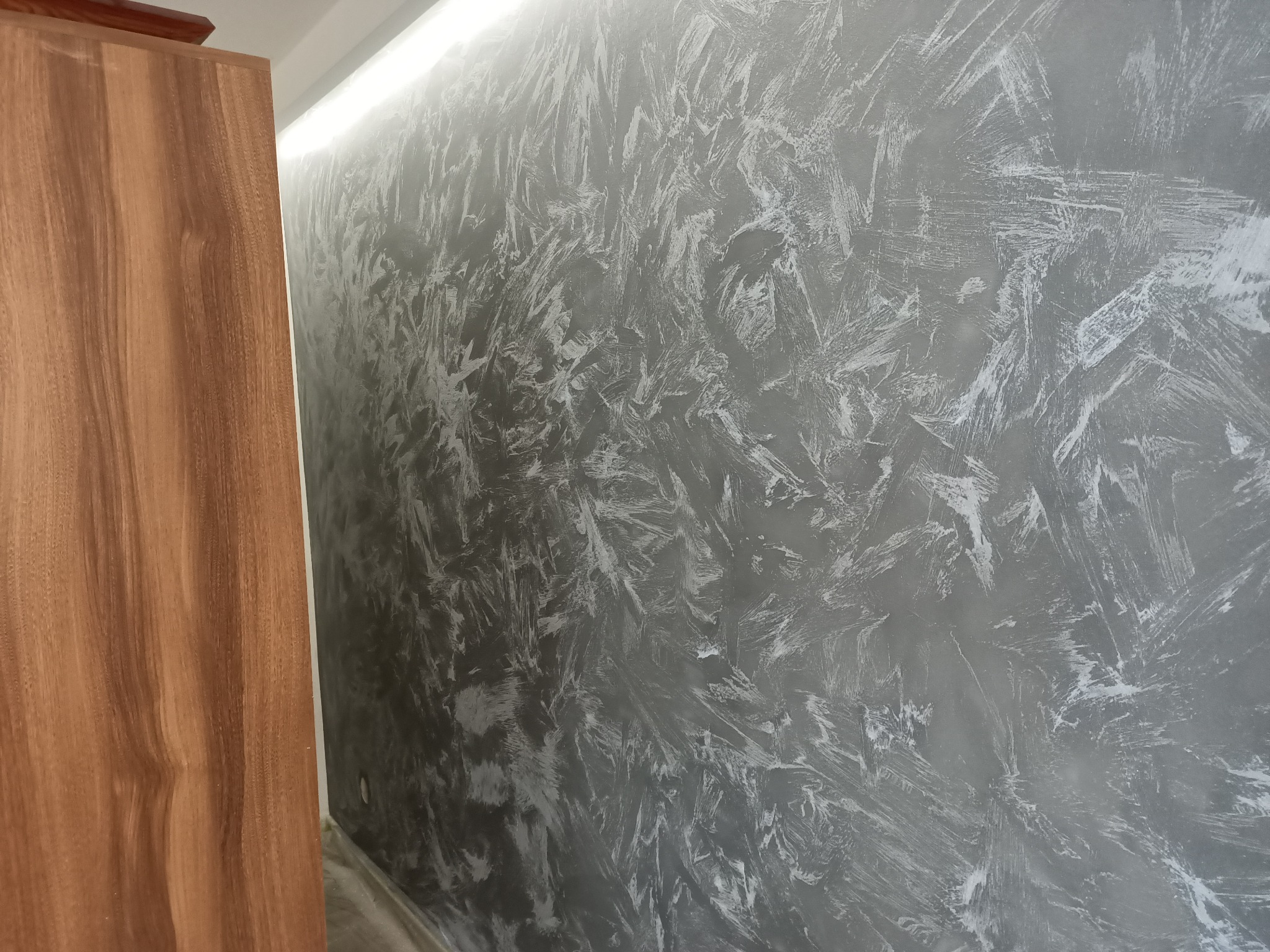 Szara ściana z dekoracyjnym efektem malowania, widoczny fragment drewnianej szafy po lewej stronie.