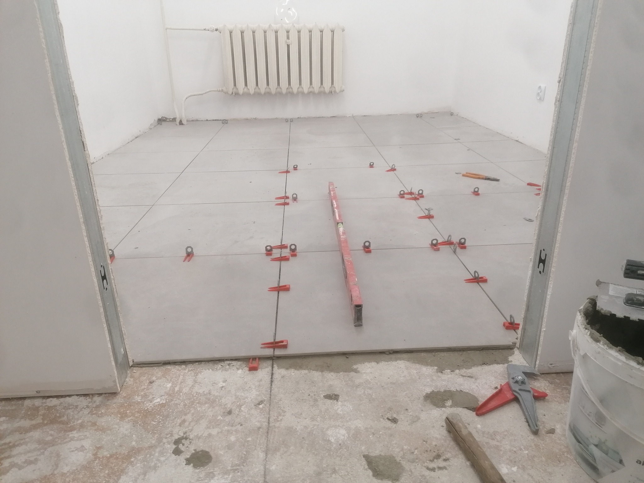 Podłoga w trakcie układania płytek, widoczne krzyżyki dystansowe, poziomica oraz narzędzia, ściany przygotowane do dalszych prac wykończeniowych, grzejnik na ścianie.