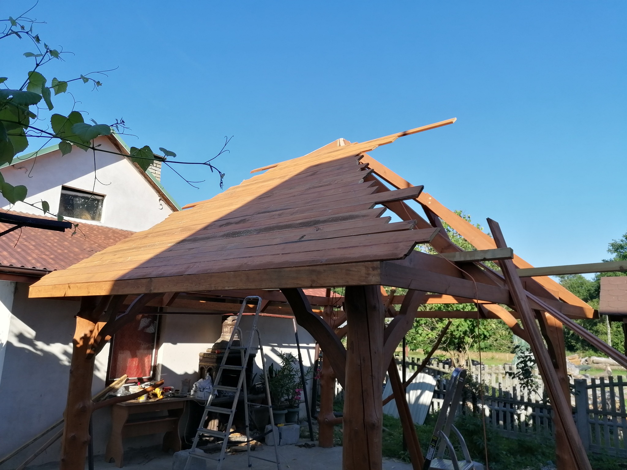 Drewniana konstrukcja z częściowo ułożonym dachem z desek, widoczne narzędzia i drabina, w tle dom i błękitne niebo.